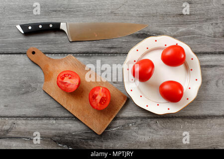 Table top view on three tomatoes on white porcelain plate with red dots, cutting board with tomato cut to half next to it, chef knife also laying on t Stock Photo