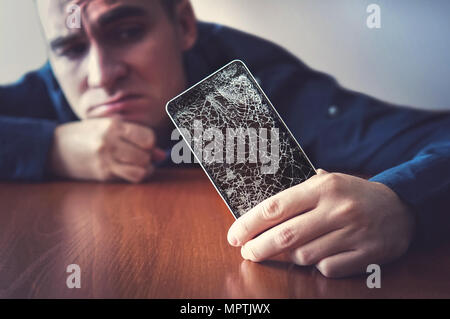 Hands holding a mobile phone with a broken screen over the wooden surface. A man looks at his broken phone lying on a wooden table with a sad look. gu Stock Photo