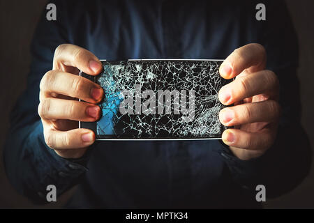 Broken phone screen in hand in dark background. Guy breaks a black modern frameless phone to which the screen cracks Stock Photo