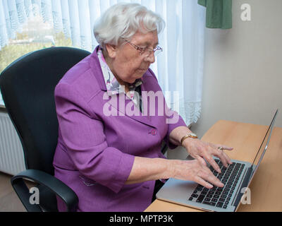 A 95-year-old woman is typing on a laptop keyboard while sitting at the desk Stock Photo
