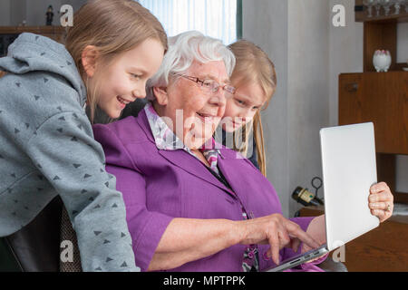 A 95 year old woman is holding a laptop  and showing her grandchildren something funny. She is sitting on a armchair in her living room and they are   Stock Photo