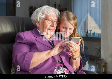 A 95 year old woman is holding a smartphone and showing her grandchild something funny. She is sitting on a armchair in her living room and they both  Stock Photo
