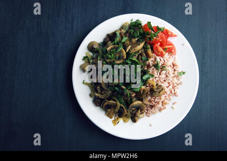 Red rice with mushrooms and cherry tomatoes. Vegan dish. European cuisine. Simple healthy lunch on the black background. Vegetarian rice dish on the w Stock Photo