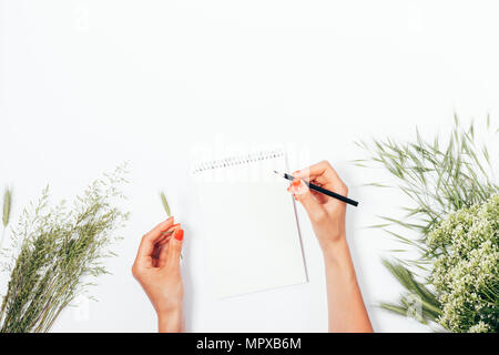 Woman writes on blank notebook page among bouquets of wild summer flowers and spikelets, top view. Flat lay festive composition female hands with red  Stock Photo