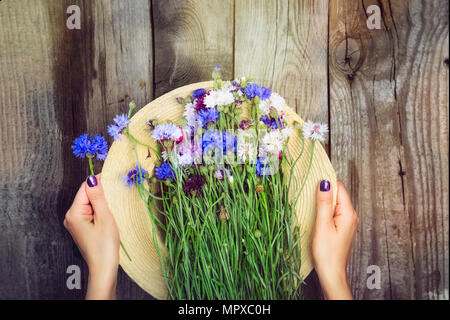 Top view woman's hands holding straw hat and romantic wild-flower bunch bouquet on the wooden vintage rustic background. Countryside lifestyle or summer relaxation concept. Text space. Stock Photo