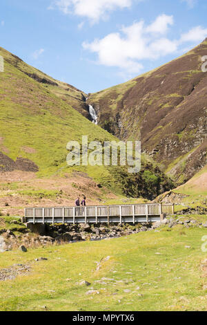 Two walkers cross the footbridge over the Tail Burn beneath The Grey Mare's Tail waterfall, near Moffat, Dumfries & Galloway, Scotland, UK Stock Photo