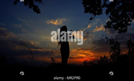 Dark female silhouette doing yoga and meditating in evening sky background. Concept of relaxing before sleeping and asanas. Stock Photo