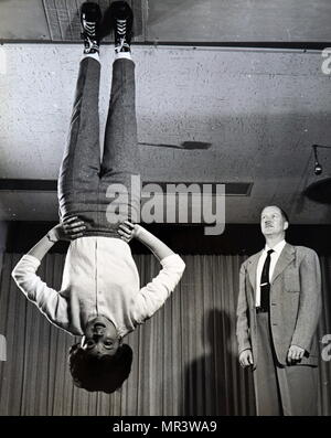Photograph depicting Nancy Wendler demonstrating zero gravity shoes for their developer, John F. Heard. The soles of the shoes are covered with minute nylon hooks that engage with a carpeted surface to create the fastening effect. The holding power of the material is great enough to suspend the weight of an average size person even with the earth's gravity pulling. In space, relatively small amounts of the hook material would be required to keep an astronaut walking properly. Dated 20th century