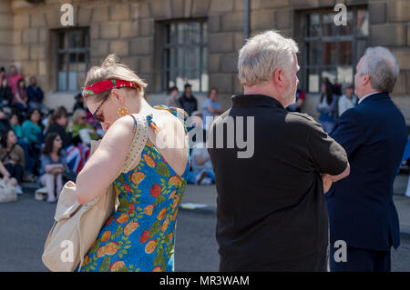 Young woman seen searching through her handbag, seen wearing a fruit themed summer dress. Seen next to a middle aged man, watching a public festival. Stock Photo