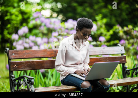African-American female with the laptop is sitting on the wooden bench. cheerful cute black girl on the park bench using the netbook. Stock Photo