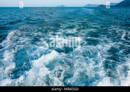 Wave trace with white foam on sea water surface behind of fast moving motorboat or yacht. Small islands in haze on horizon. Blue sky before sunset. Stock Photo
