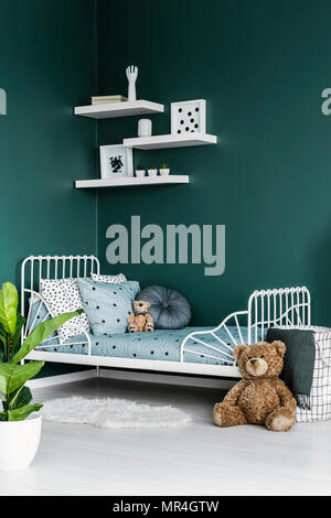 Teddy bear toy by a white twin bed in a dark green room interior for a child with white decor and a plant Stock Photo