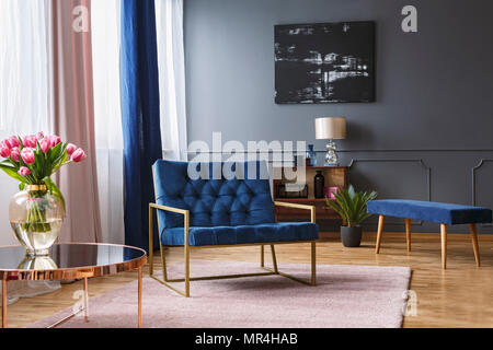 Real photo of a blue, wide chair standing on a rug in a spacious living room interior with grey walls and wooden floor next to a shelf and a table Stock Photo