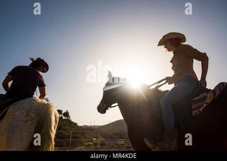 couple of modern cowboys man and woman ride two horses outdoor with sunflare and backlight. mountains and wind mill in background. nice young on vacat Stock Photo