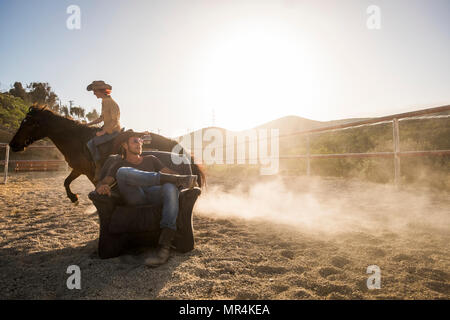 couple of beautiful man and woman together. she rides a horse making dust with speed and he stays sit down on the chair in the middle of the track Stock Photo