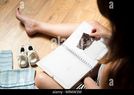 Woman placing baby's sonogram into baby's first year memory book. Baby clothes and sneakers laying on the floor Stock Photo