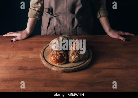 partial view of woman standing at table with loafs of bread on wooden cutting board Stock Photo