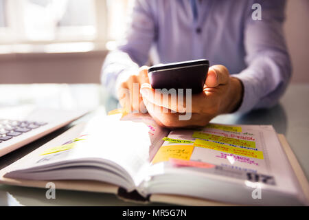 Businessperson Using Mobile Phone With Diary On Desk Stock Photo