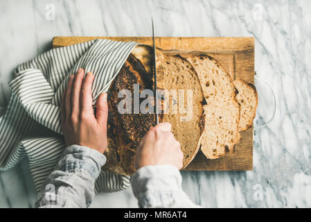 Flat-lay of female hands cutting freshly baked sourdough bread into pieces on rustic wooden chopping board over light grey marble background, top view Stock Photo