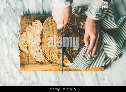 Flat-lay of woman's hands cutting freshly baked sourdough bread with knife into pieces on rustic wooden chopping board over light grey marble backgrou Stock Photo