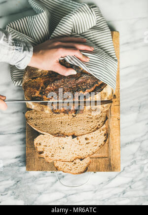 Flat-lay of woman's hands cutting freshly baked sourdough bread with knife into pieces on rustic wooden chopping board over light grey marble backgrou Stock Photo