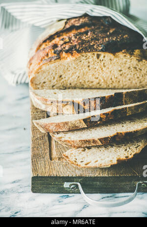 Flat-lay of freshly baked sourdough bread cut into pieces on rustic wooden chopping board over light grey marble background, selective focus, vertical Stock Photo