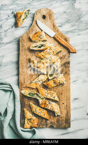 Flat-lay of fresh Turkish borek roll cut in slices slices with spinach, feta cheese, black cumin seeds on wooden board over grey marble background, to Stock Photo