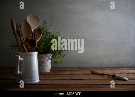 A still life image of a rustic, wood kitchen counter with wooden spoons in a porcelain vessel and a lovely, tangled oregano plant in the background. Stock Photo