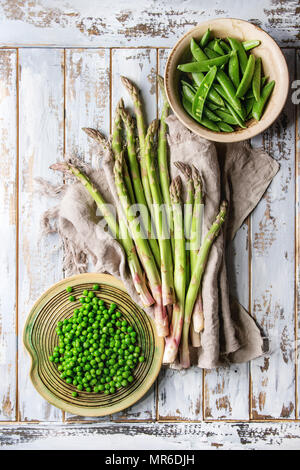 Variety of raw uncooked organic young green vegetables asparagus, peas, pod pea in ceramic plates on linen cloth over white wooden plank background. T Stock Photo