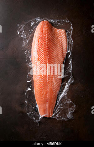 Whole raw uncooked salmon fillet on food film with chef's knife over dark brown texture background. Top view, space. Stock Photo