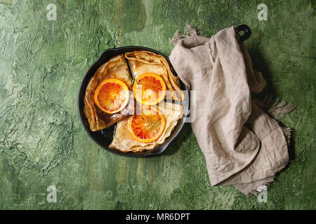 Homemade crepes pancakes served in cast-iron pan with bloody oranges and rosemary syrup with sliced sicilian red oranges over green texture background Stock Photo