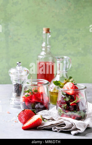 Beetroot and strawberry salad served with arugula and nuts in glass jars with cloth, pepper and bottles of fruit ocet and olive oil over grey table wi Stock Photo