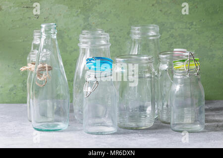 Variety of different shape empty opened glass bottles with and without lids standing on grey table with green wall as background. Stock Photo