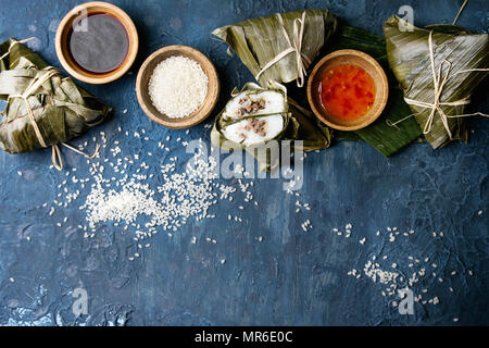 Asian rice piramidal steamed dumplings from rice tapioca flour with meat filling in banana leaves. Ingredients and sauces above over blue texture back Stock Photo