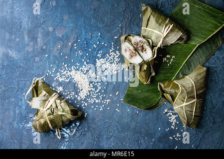 Asian rice piramidal steamed dumplings from rice tapioca flour with meat filling in banana leaves with rice above over blue texture background. Top vi Stock Photo