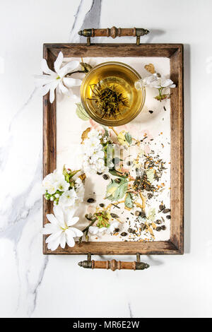 Glass cup of hot green tea on vintage tray with spring flowers white magnolia and cherry blooming branches over white marble texture background. Top v Stock Photo