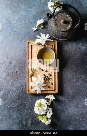 Traditional ceramic cup of hot green tea on wooden board with black iron teapot, spring flowers white magnolia and cherry blooming branches over dark