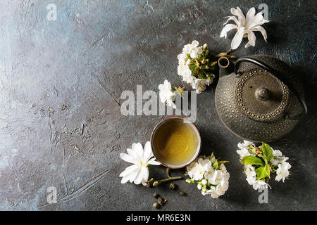 Traditional ceramic cup of hot green tea with black iron teapot, spring flowers white magnolia and cherry blooming branches over dark blue texture bac Stock Photo