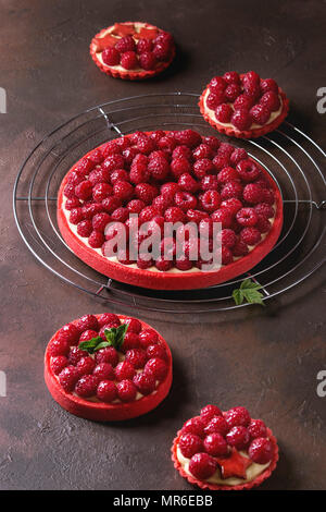 Variety of red raspberry shortbread tarts and tartlets with lemon custard and glazed fresh raspberries served on cooling rack over dark brown texture Stock Photo