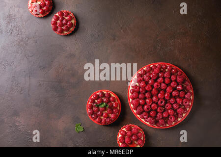 Variety of red raspberry shortbread tarts and tartlets with lemon custard and glazed fresh raspberries over dark brown texture background. Top view, s Stock Photo