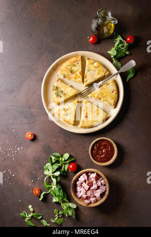 Sliced spanish potato omelette tortilla with bacon served in ceramic plate with sauce and ingredients above over dark brown texture background. Top vi Stock Photo