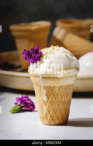 Homemade vanilla ice cream in small waffle cup served with purple edible flowers and metal spoon in ceramic plate on white marble kitchen table. Stock Photo