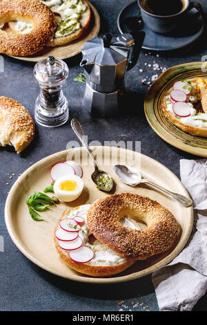 Variety of homemade bagels with sesame seeds, cream cheese, pesto sauce, eggs, radish, herbs served on ceramic plate with ingredients and coffee above Stock Photo