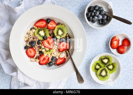 Bowl of muesli, sliced kiwifruit, strawberries and blueberries with a spoon and small bowls of fruit. Stock Photo