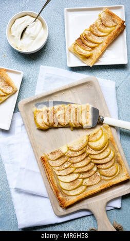A rectangular apple tart on a wooden serving board cut in pieces being served onto dessert plates. Stock Photo