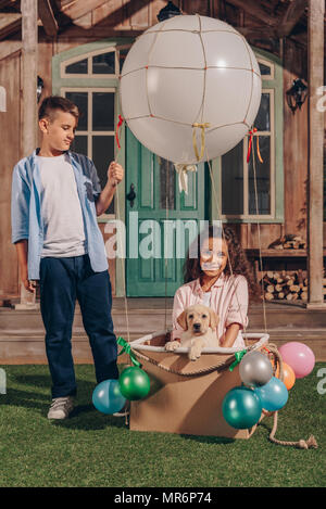 african american girl with labrador puppy sitting in air balloon box while boy standing near by Stock Photo