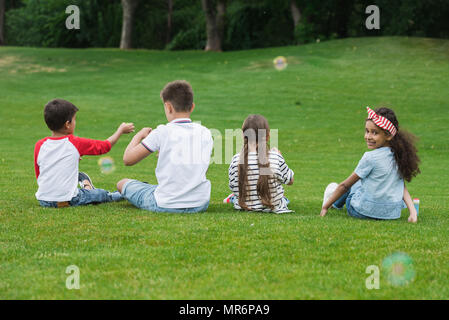 Back view of cute multiethnic kids playing with soap bubbles while sitting on green grass in park Stock Photo
