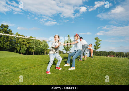 Multiethnic group of kids playing tug of war on green grass in park Stock Photo