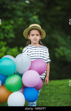 Cute little girl in straw hat holding bunch of colorful balloons and looking at camera Stock Photo