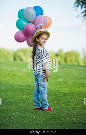 Adorable little girl in straw hat holding bunch of colorful balloons while standing in park Stock Photo